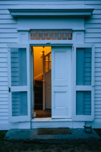 At a white house tinged blue by twilight, the door is open to a small stairhall with a warm yellow light.