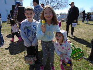 Three young siblings smile while holding baskets and a handmade chick craft