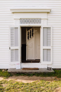 Doorway with a transom window and double screen doors opens to a stairhall with shaped balusters and paneled woodwork