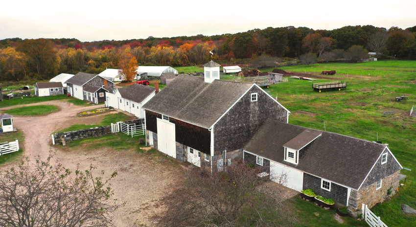 Aerial of row of shingled barns and white outbuildings with sandy barnyard in front, green fields in back, and autumn trees in background
