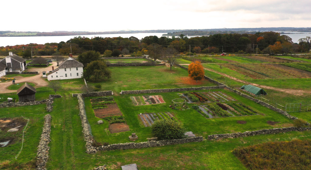 Aerial of garden, lawn, barnyard, and planting fields with bay and islands beyond. White farmhouse at center left.