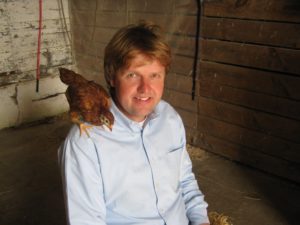 Adam Lowe sits in a barn and smiles as a Rhode Island Red chicken perches on his shoulder.