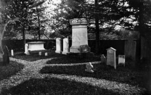 In a burial ground bordered by trees and walls, white tombstones of varying shapes are connected by light-colored paths.