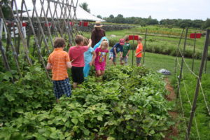 Eight young children and one adult near vegetable garden rows of bush beans, staked pole beans, and carrots