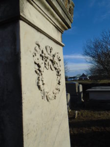 An aged tall gravestone of white marble carved with a round oak garland in the foreground with farm buildings in the distance