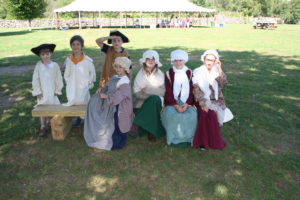 In the shade of a tree, seven children are dressed in reproduction colonial clothing.