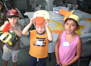 Three young children hold construction paper hats that look like ducks. Materials for making the crafts are on a table behind them.