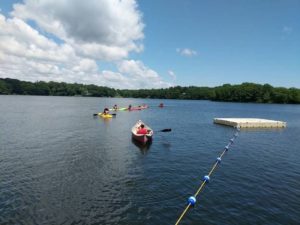 Under blue sky and fluffy clouds, kayakers paddle out on blue water near a floating rope and raft