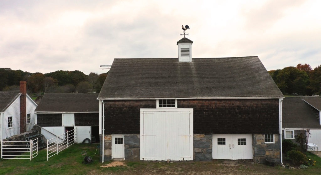 Barn with a tall stone foundation, shingled siding and roof, large white doors and small windows, and a cupola with a rooster weathervane.