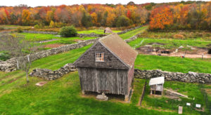 Wood-slat shed with a square window in the gable end, slightly canted sides, and set on stone pillars. Bright autumn trees behind.