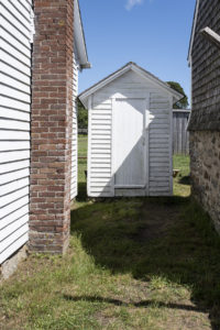 Small white clapboarded shed with a closed door in the narrow alley between two larger outbuildings