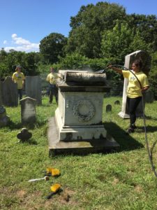 Brushes on the ground, two young people with yellow shirts stand in the burial ground while one sprays a tombstone with a hose.
