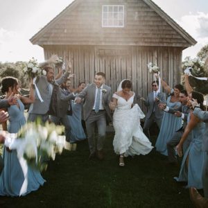 From a weathered wood building, a bride and groom run down the lawn between two rows of cheering bridesmaids and groomsmen.