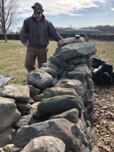 Bearded man wearing a hat stands next to a partially dismantled stone wall
