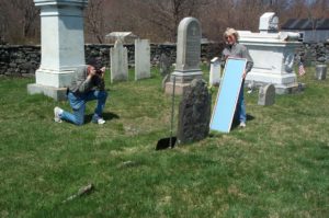 A man kneels in front of a slate headstone with a camera while a woman holds a full-length mirror to shine light on the stone.