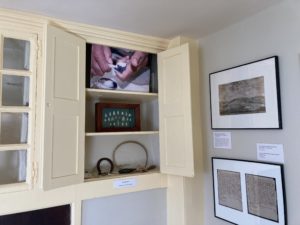 Shelves with stone projectile points, a picture of hands making wampum, and small items. Two framed pictures on the wall.