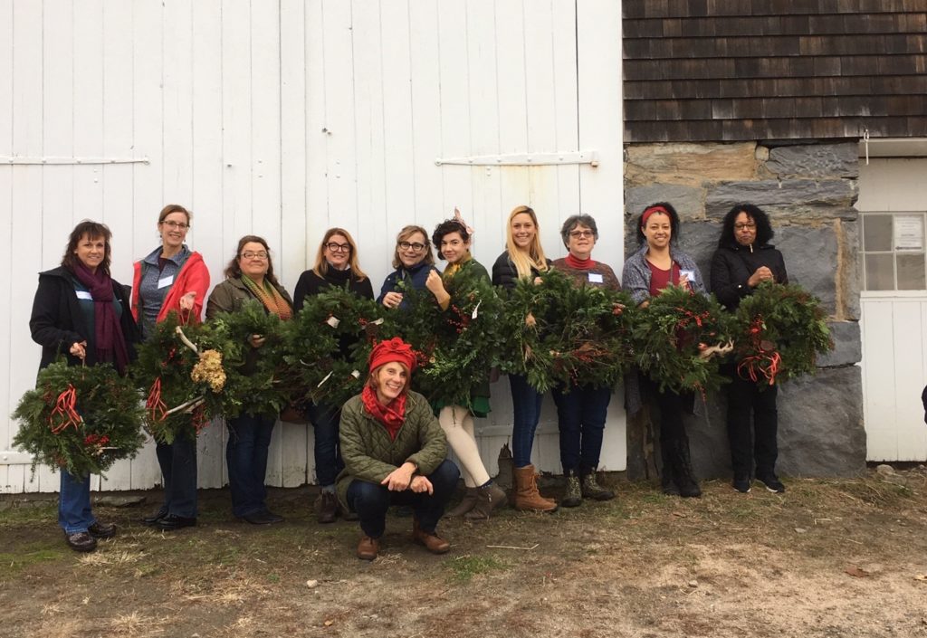 At a large barn, ten women hold up round green wreaths with red bows and decorations. Their smiling teacher crouches in front.