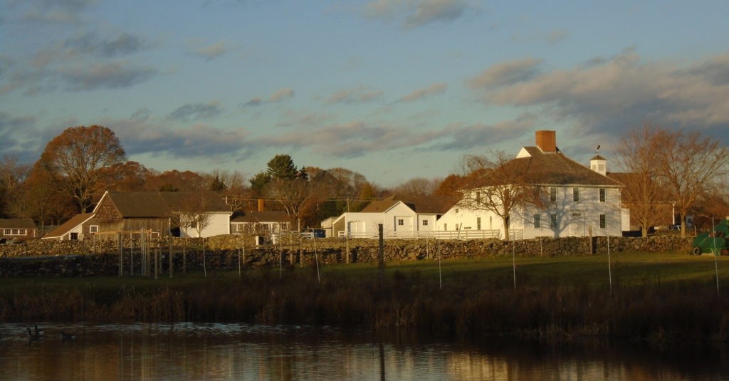 A long view of a white house, carriage house, and shingled agricultural buildings is reflected in still water with dark clouds in the sky.