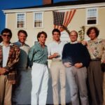 Seven men stand in front of the yellow Bowman House with an American flag in the background